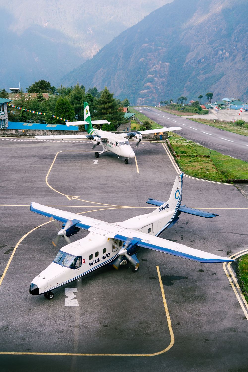 Two small white passenger aircraft parked on mountain airstrip with dramatic peaks in background.
