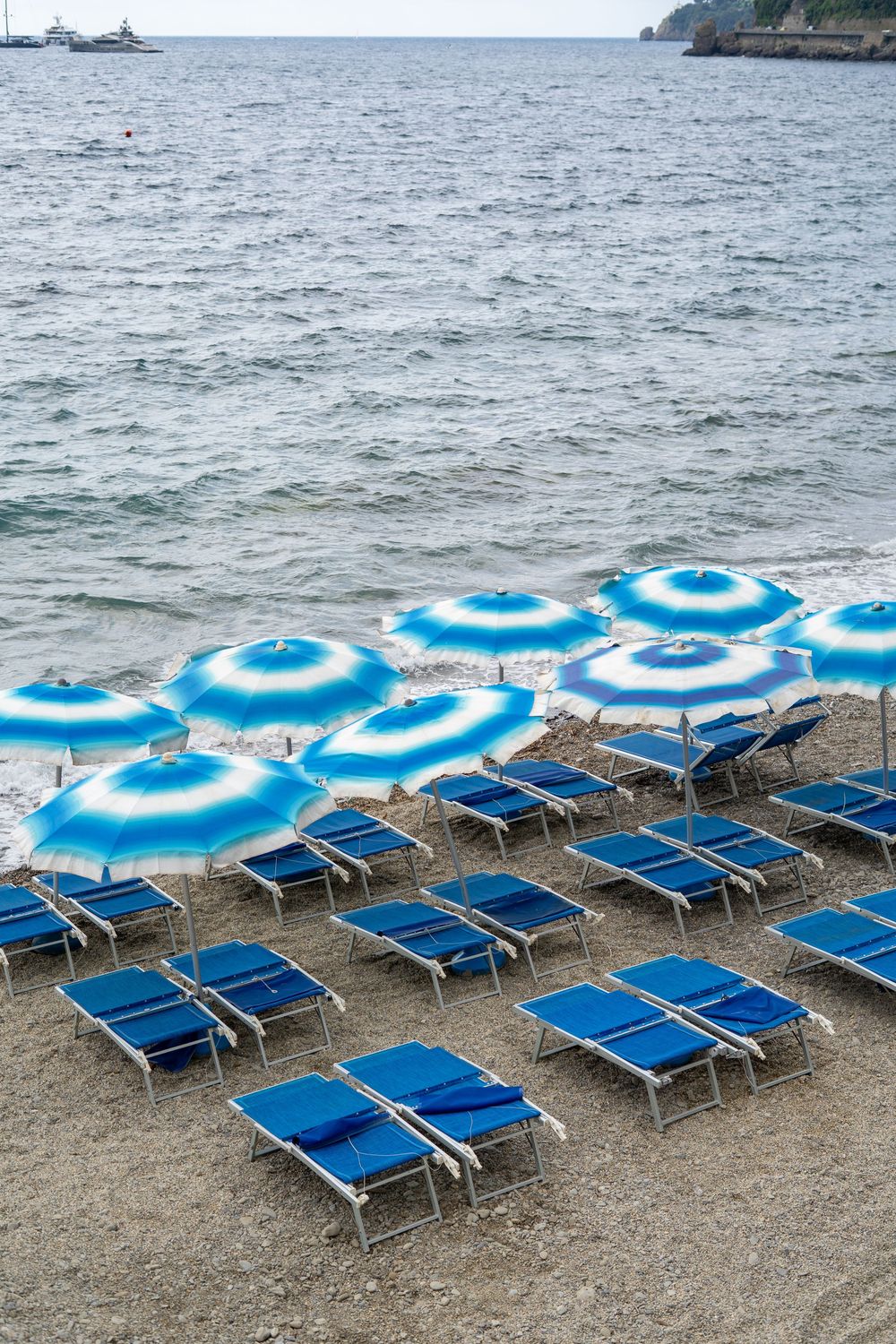 Blue and white striped beach umbrellas and loungers lined up on a rocky Mediterranean beach.