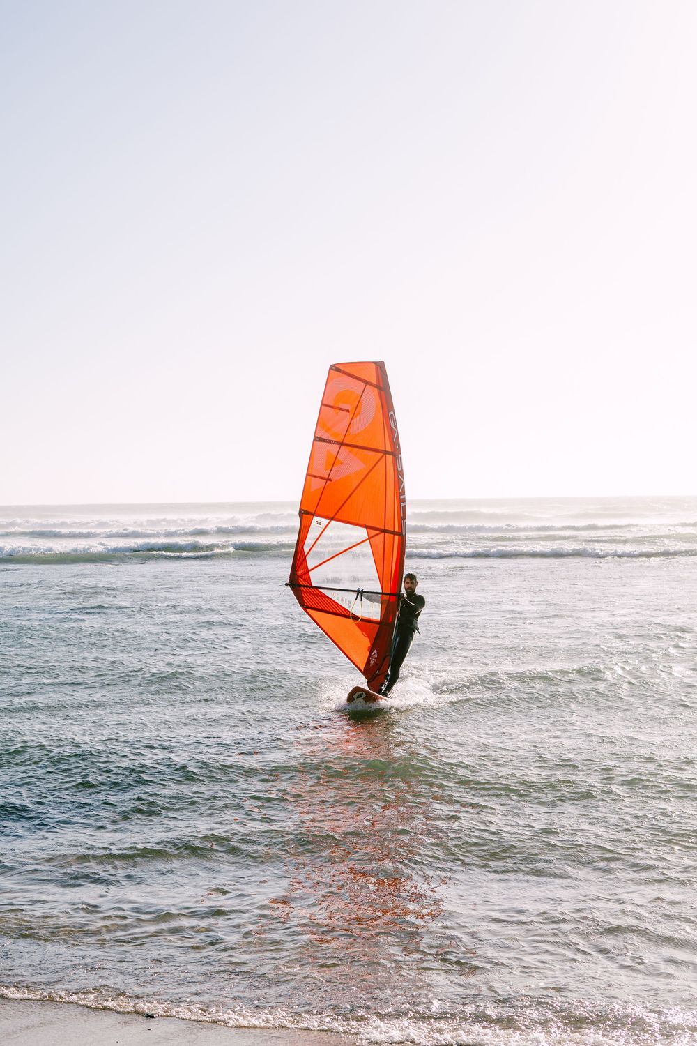 Windsurfer with orange sail riding waves at beach during sunset.