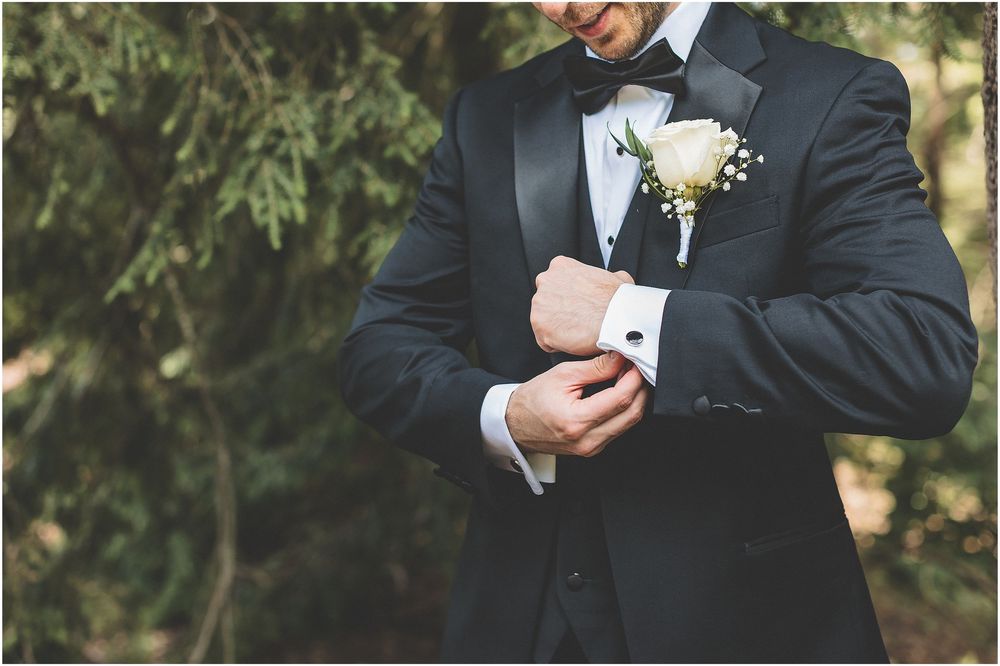 Close-up view of groom in black tuxedo adjusting cufflink with white boutonniere on lapel.
