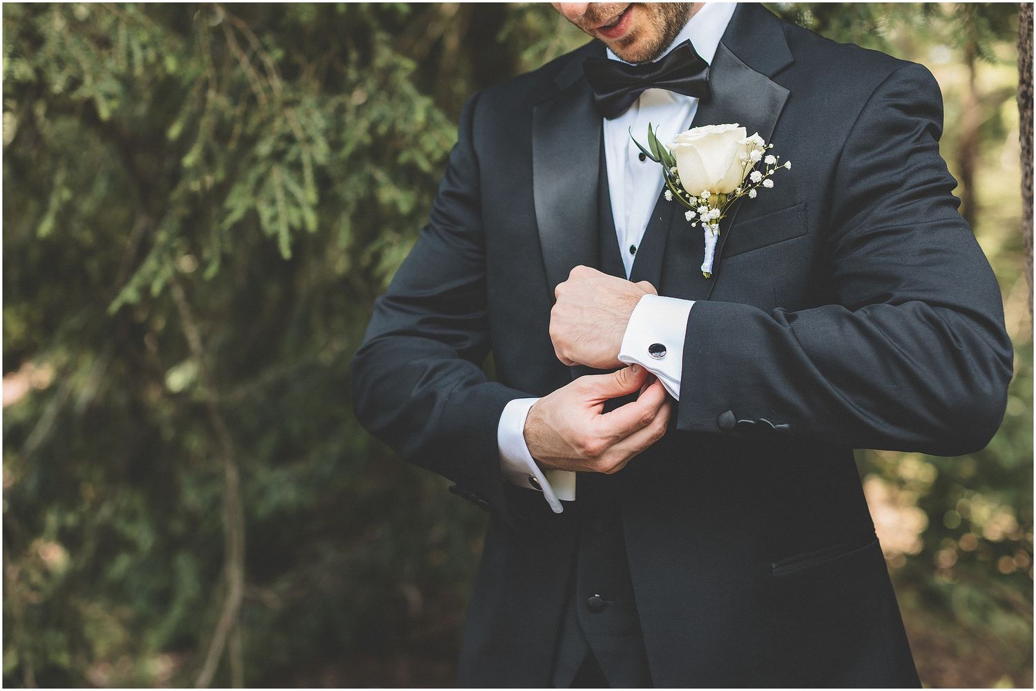Close-up view of groom in black tuxedo adjusting cufflink with white boutonniere on lapel.