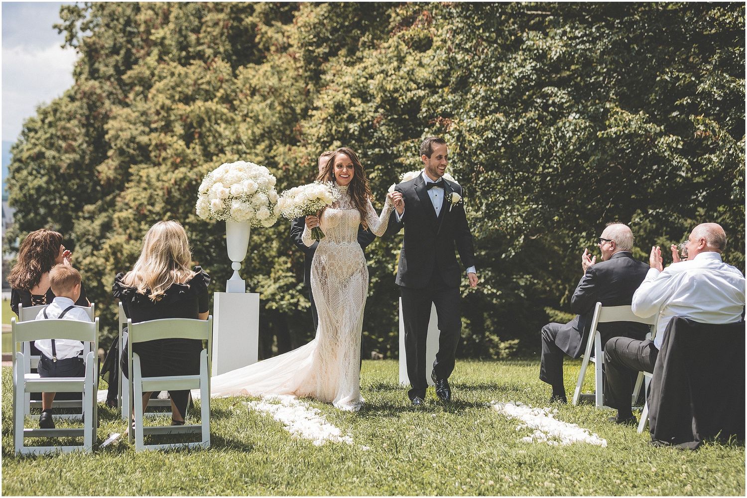 Small wedding ceremony takes place outdoors under trees on a grassy lawn during a summer afternoon.