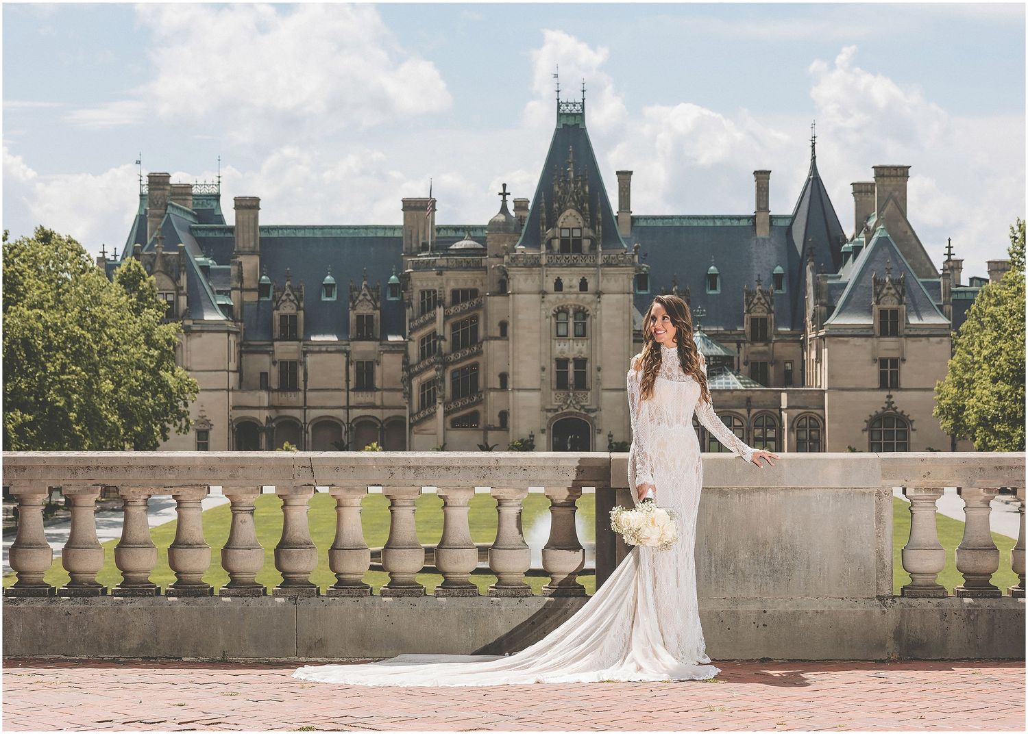 A bride in a flowing white gown walks through the grounds of an elegant stone castle estate on her wedding day.