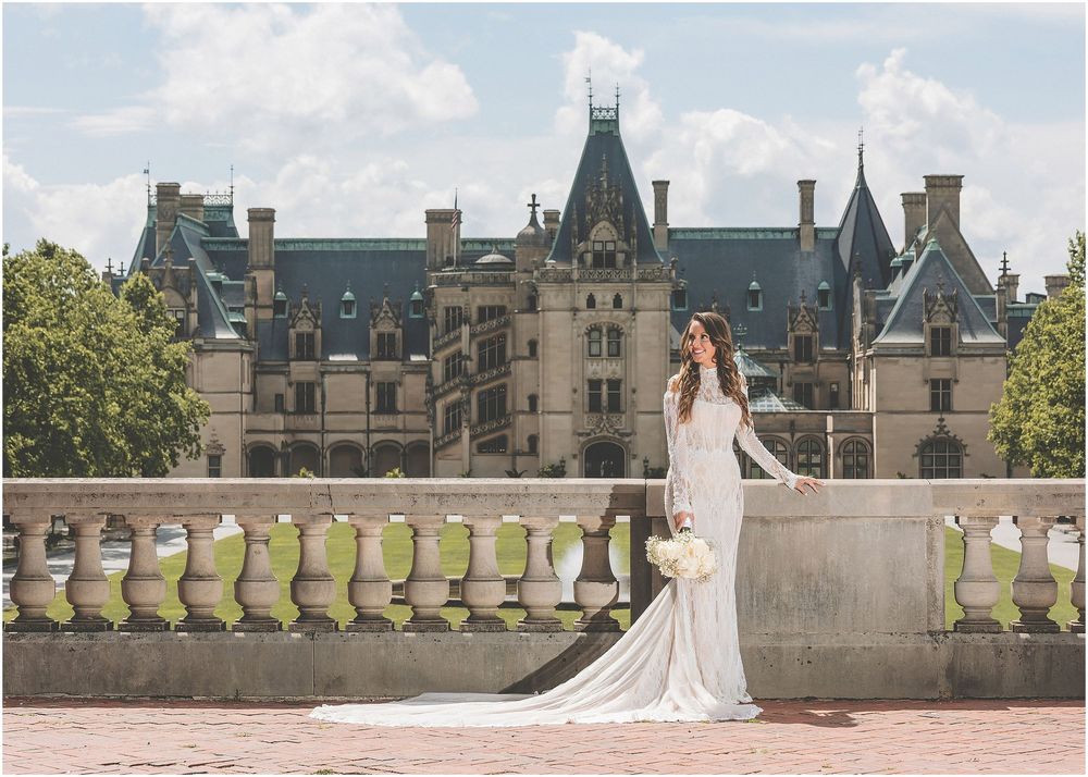 A bride in a flowing white gown walks through the grounds of an elegant stone castle estate on her wedding day.