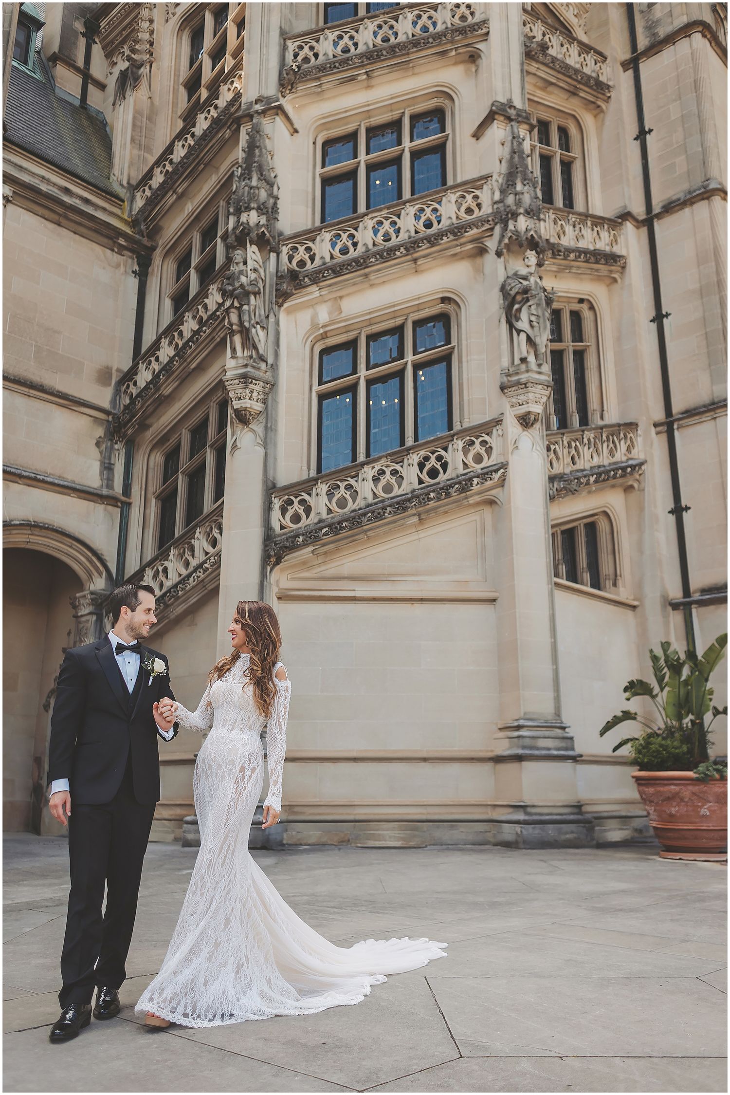 A couple in formal wedding attire poses together outside an ornate stone building with Gothic architectural details.