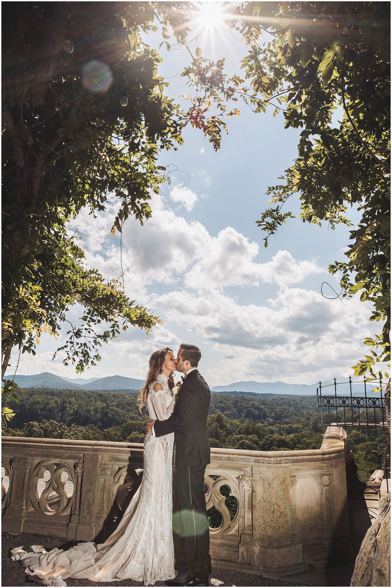 A couple shares a romantic moment on a stone balcony overlooking a scenic mountain landscape on a sunny afternoon.