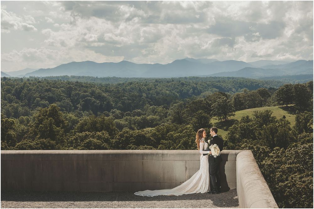 Wedding couple walks on concrete bridge overlooking scenic mountain lake and forest landscape at sunset.