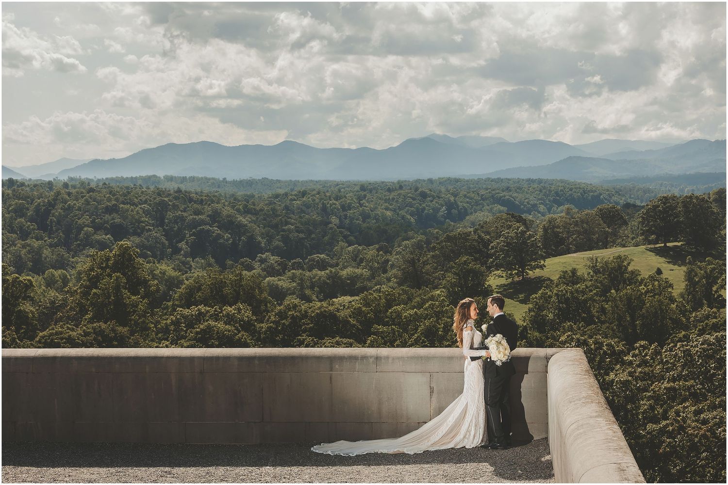 Wedding couple walks on concrete bridge overlooking scenic mountain lake and forest landscape at sunset.