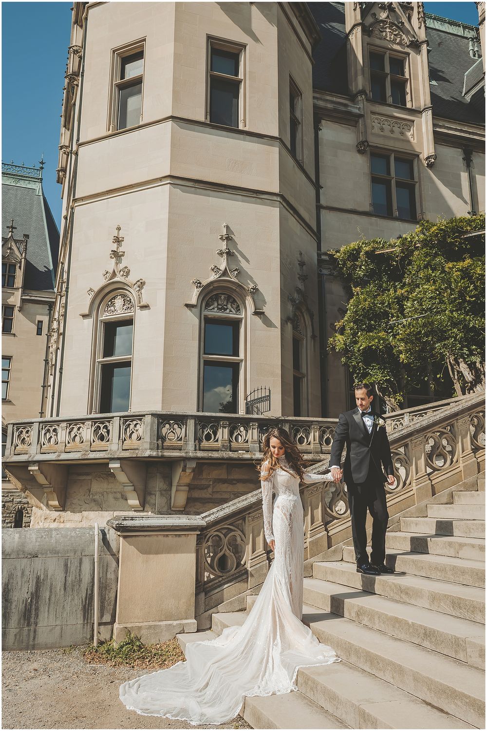 A couple in formal attire poses for photos on an elegant stone staircase with ornate railings at a historic building.