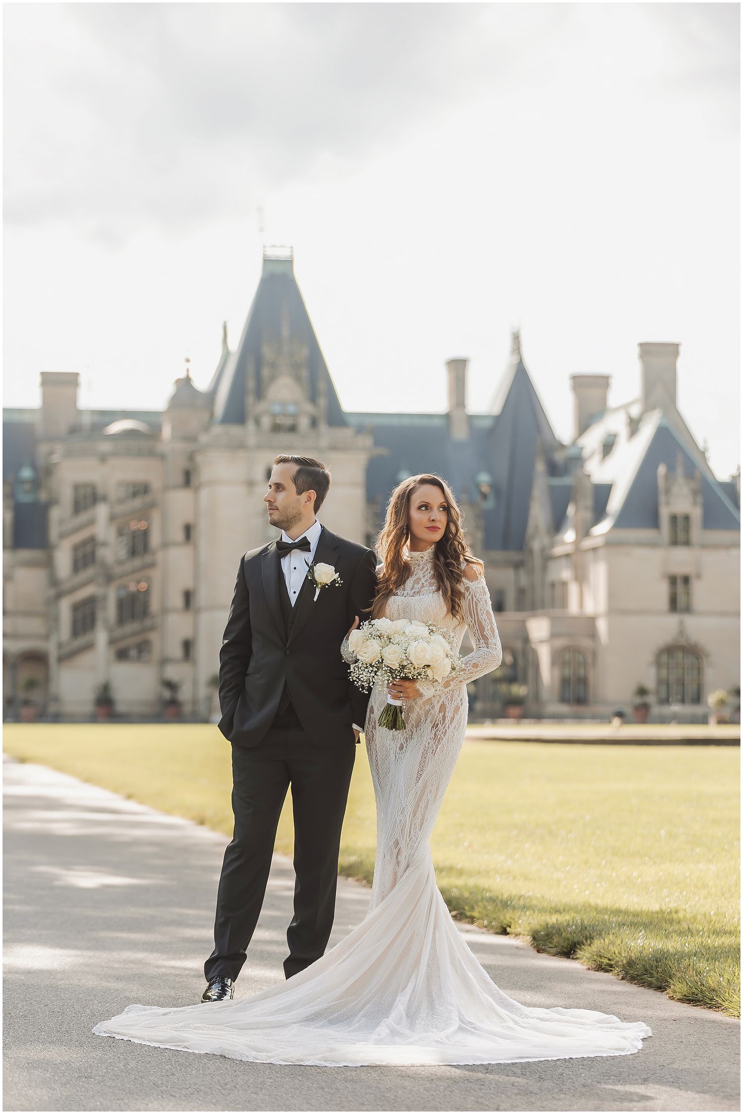 A couple poses on the grounds of an elegant estate with a Gothic stone mansion in the background.
