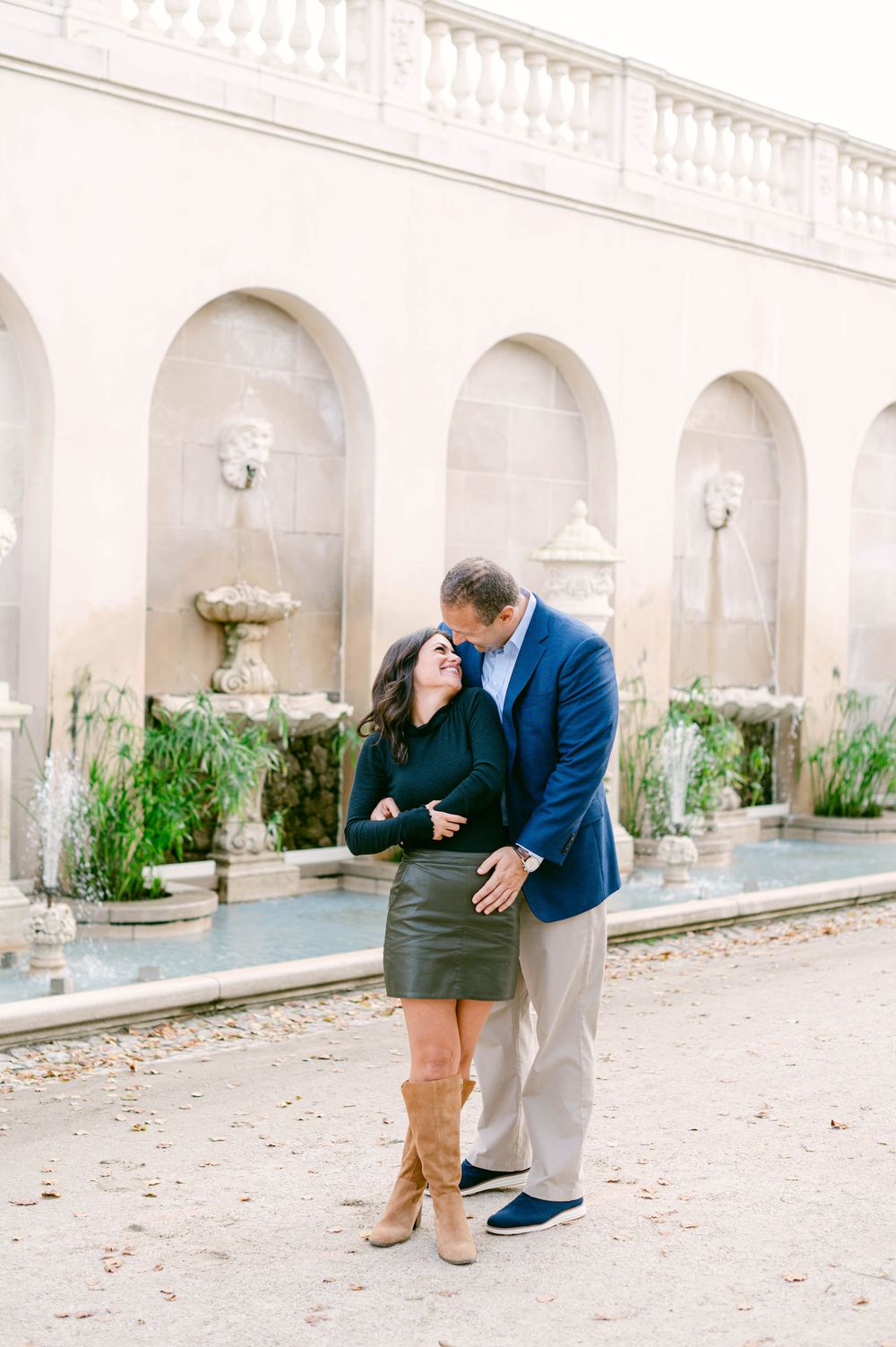 A couple shares a playful moment along an elegant white stone colonnade with fountains.