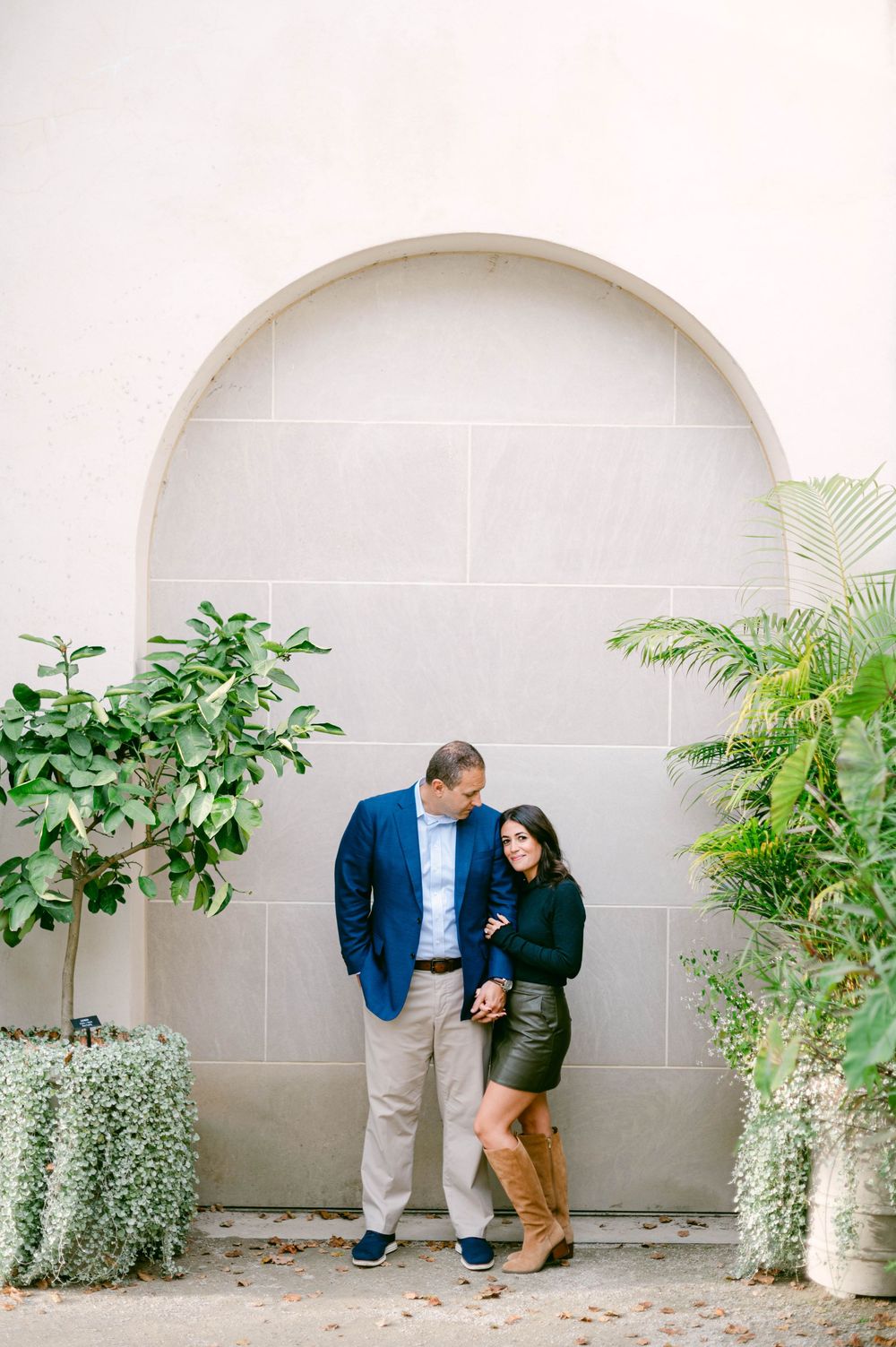 Couple standing together against arched wall with tropical plants framing the scene.