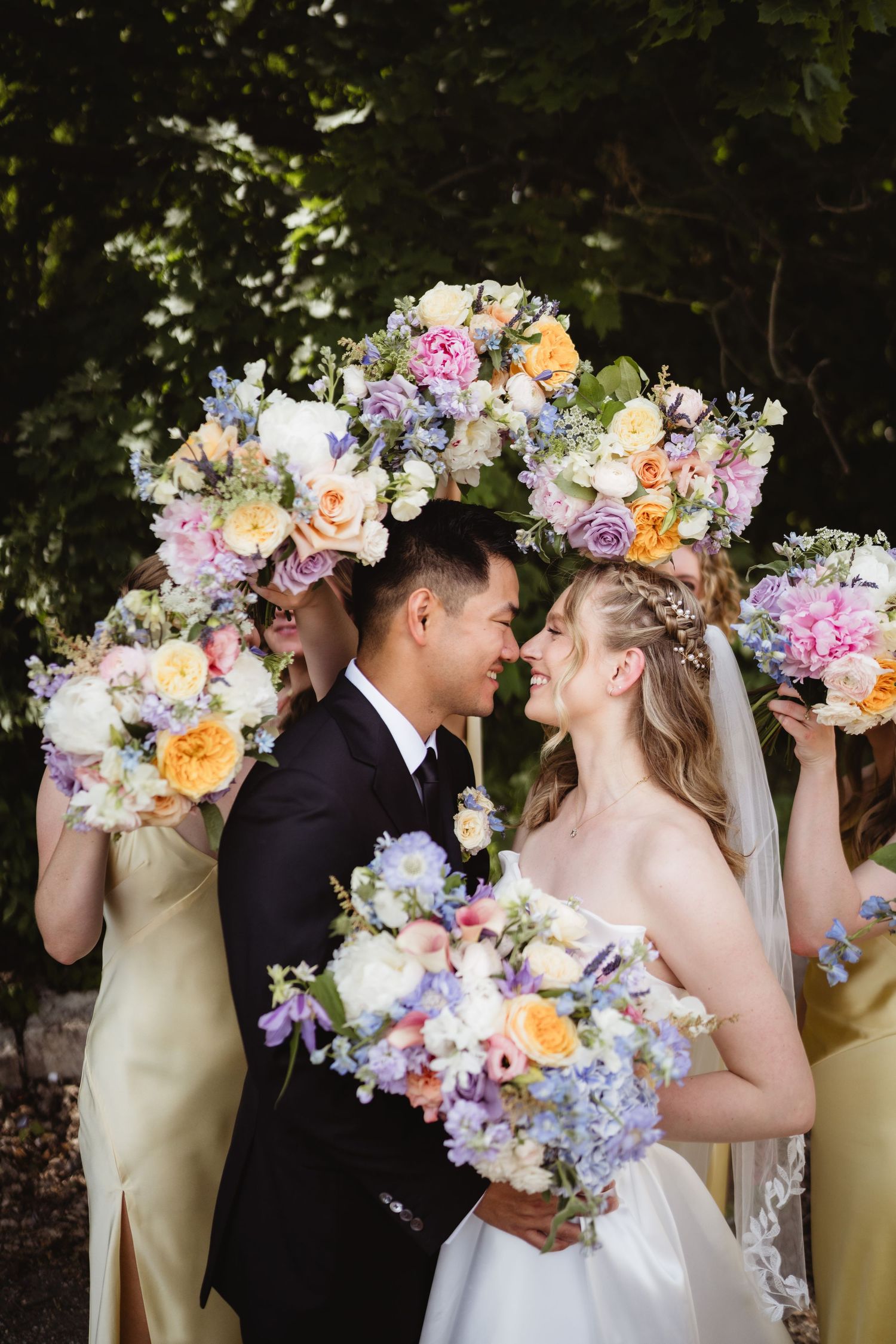 Beautiful wedding party with pastel floral arch and bouquets featuring yellow, pink, lavender and peach blooms outdoors.