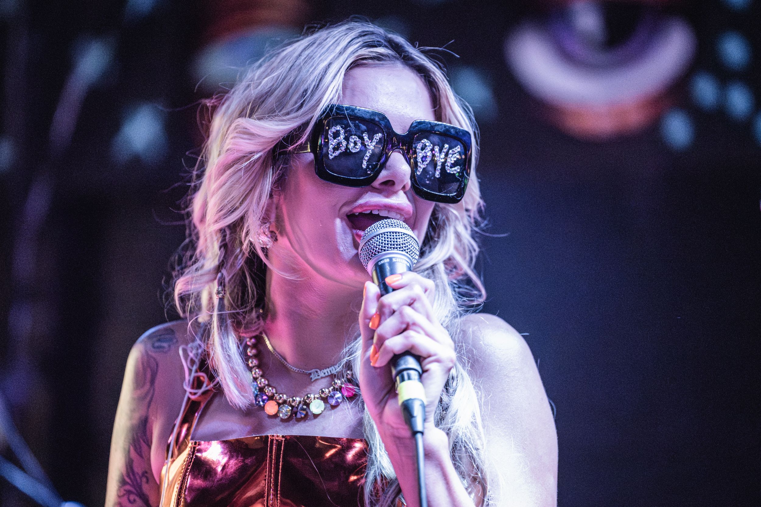 Singer wearing sunglasses with the text 'BOY BYE' performs on stage under concert lighting.