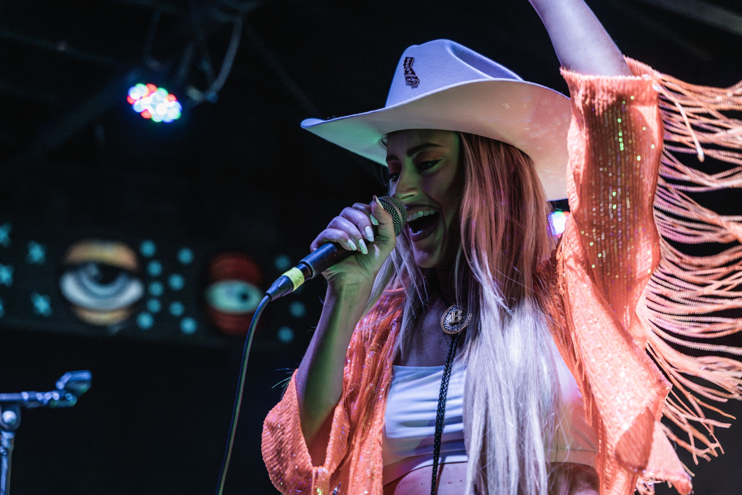 A performer in a cowboy hat singing on stage in colorful lighting with fringed sleeves on their outfit.