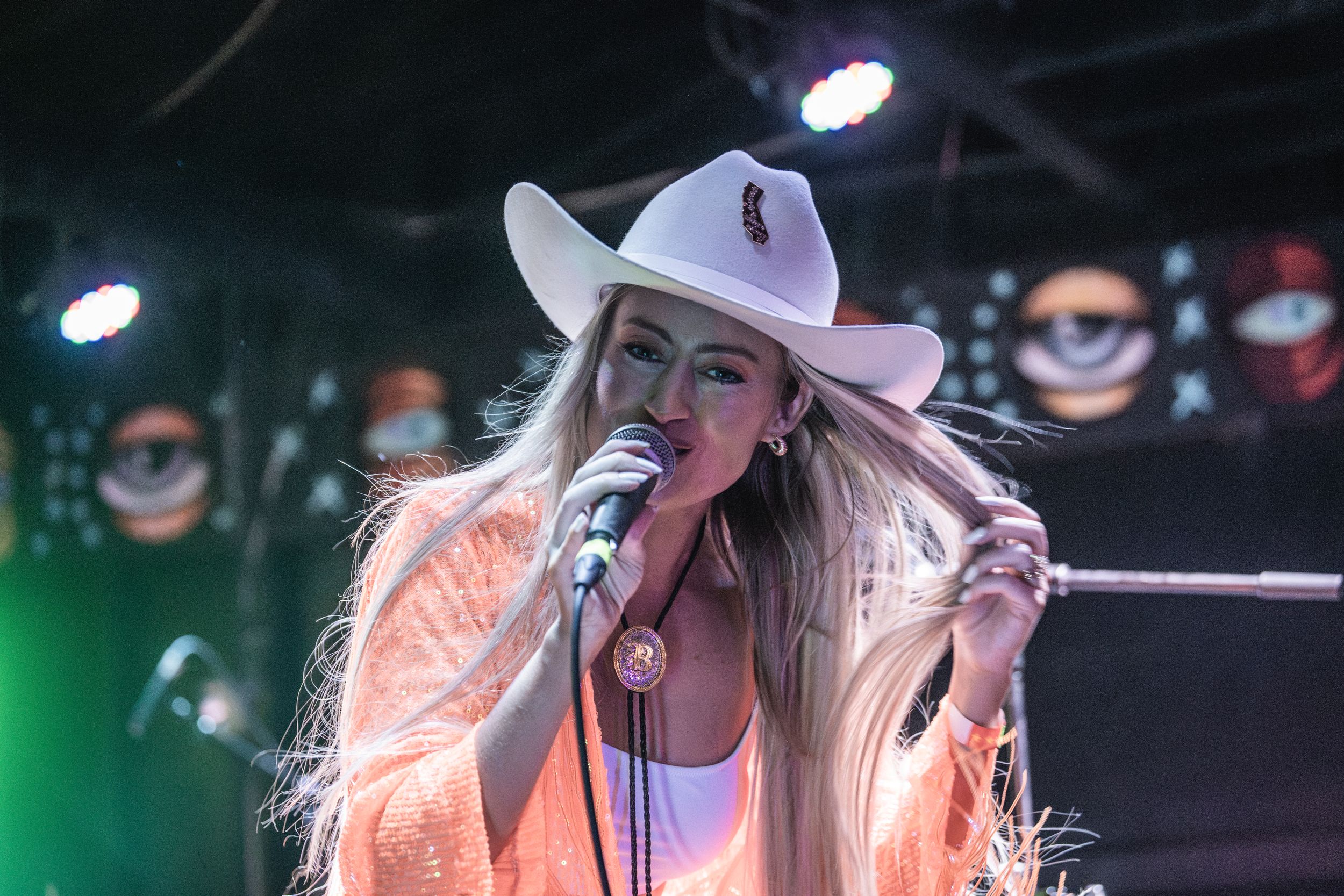 Country music performer wearing a white cowboy hat and pink sequined jacket sings with passion on a dark stage.