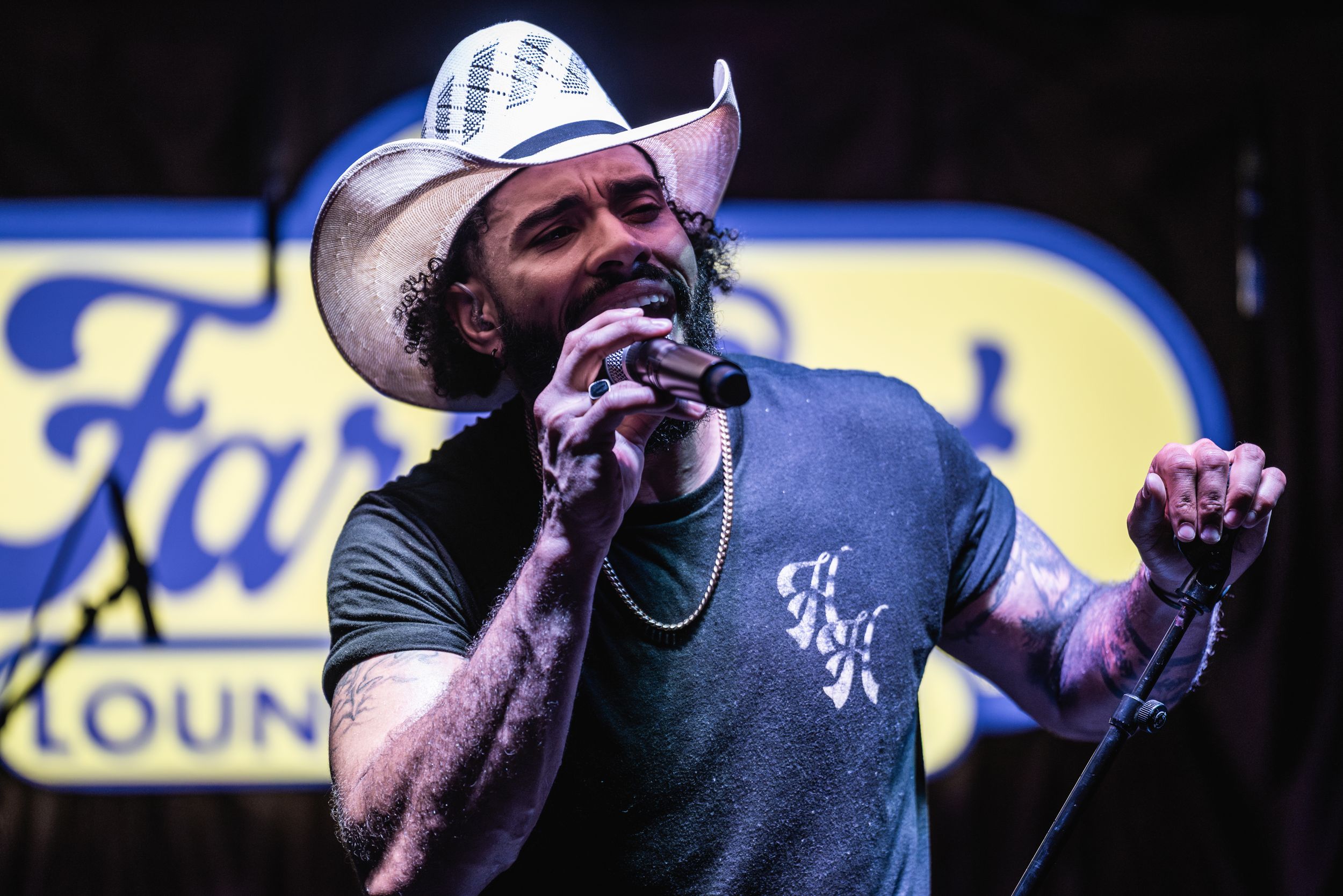 A country music performer sings passionately on stage wearing a cowboy hat and gray t-shirt with text.