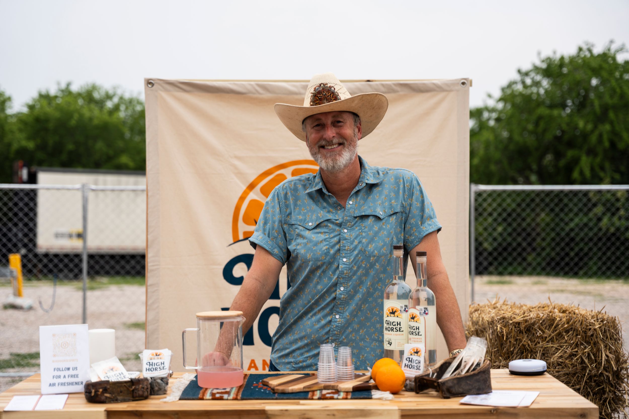 A vendor in a cowboy hat and blue shirt stands behind a display table with hay bales at an outdoor market event.