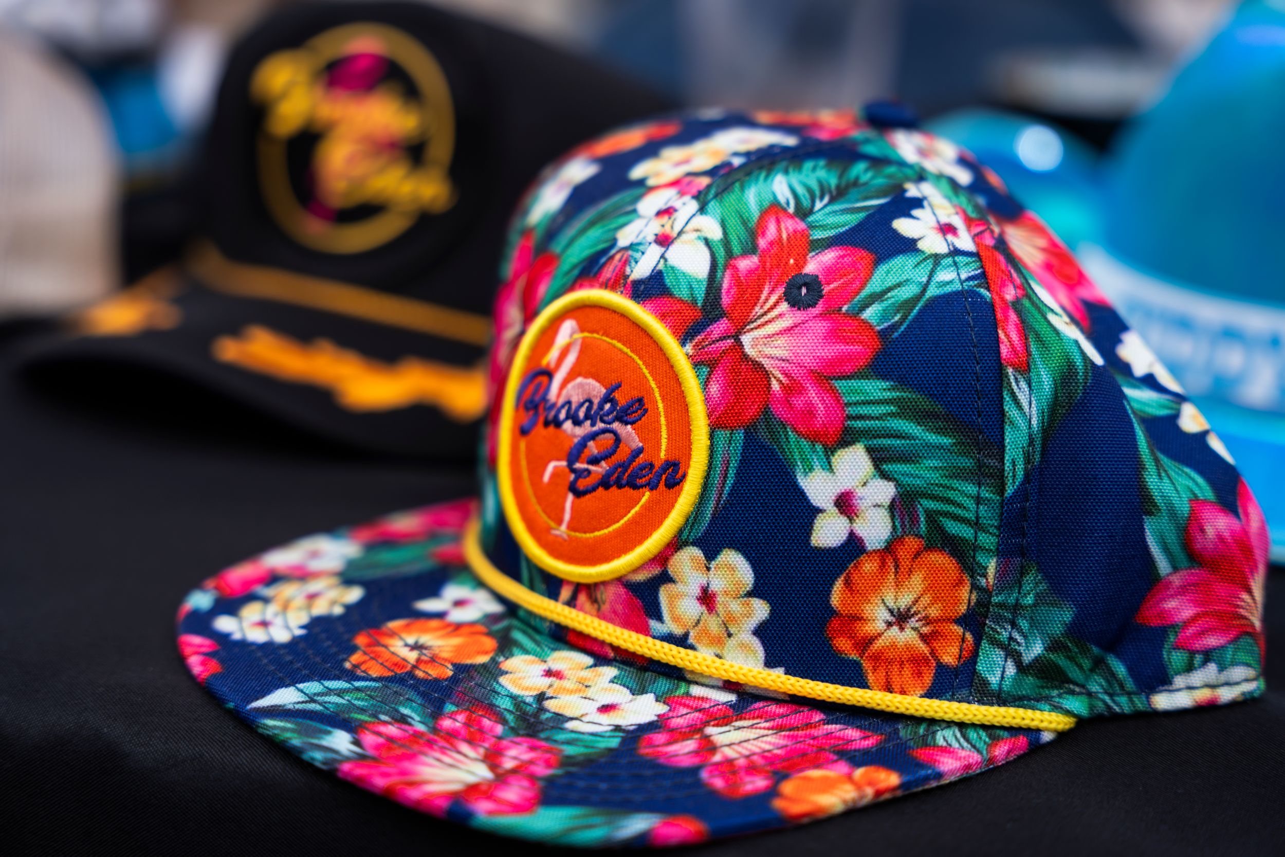 A vibrant floral print snapback hat with tropical flowers and an orange circular logo patch on navy blue background.