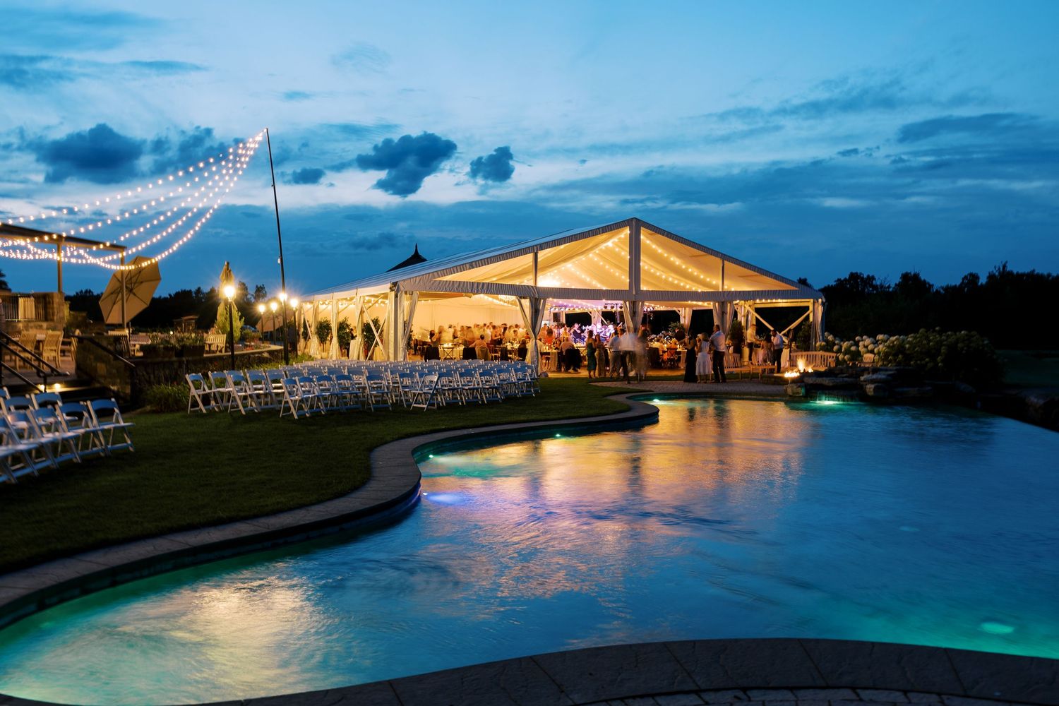 Evening wedding reception venue with illuminated tent pavilion and string lights reflecting on a glowing pool at dusk.