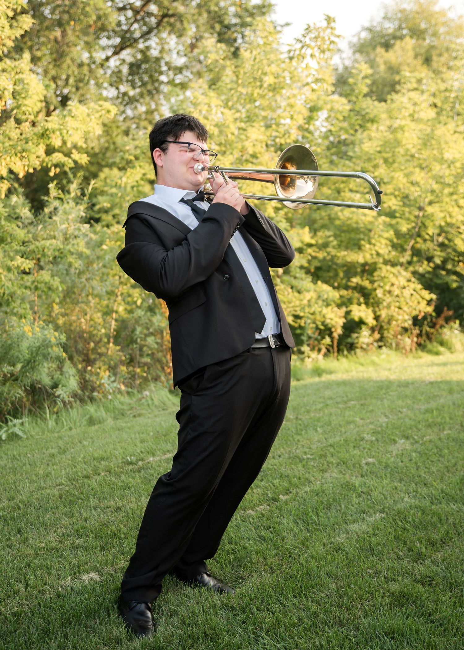 A musician in a black tuxedo performs on a trombone while leaning back dramatically on a grassy hillside.
