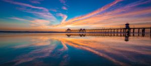 Long exposure of a coastal pier silhouetted against orange and blue sunset sky with mirror reflections in the water.