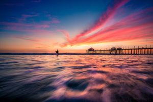 A vibrant sunset illuminates the sky in pink and blue hues above a wooden pier stretching into the calm ocean waters.