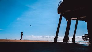 Silhouettes of beachgoers and pier structures against a bright blue sky at the coast.