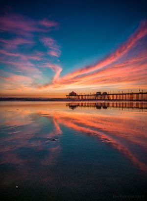 Dramatic pink and orange clouds reflect perfectly in the wet sand beneath a long oceanfront pier at sunset.