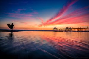 Colorful ocean sunset with pink and orange clouds reflecting on calm waters near a pier.