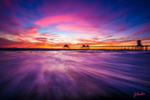 Purple and pink sunset creates dynamic reflections under a wooden pier with motion-blurred waves.