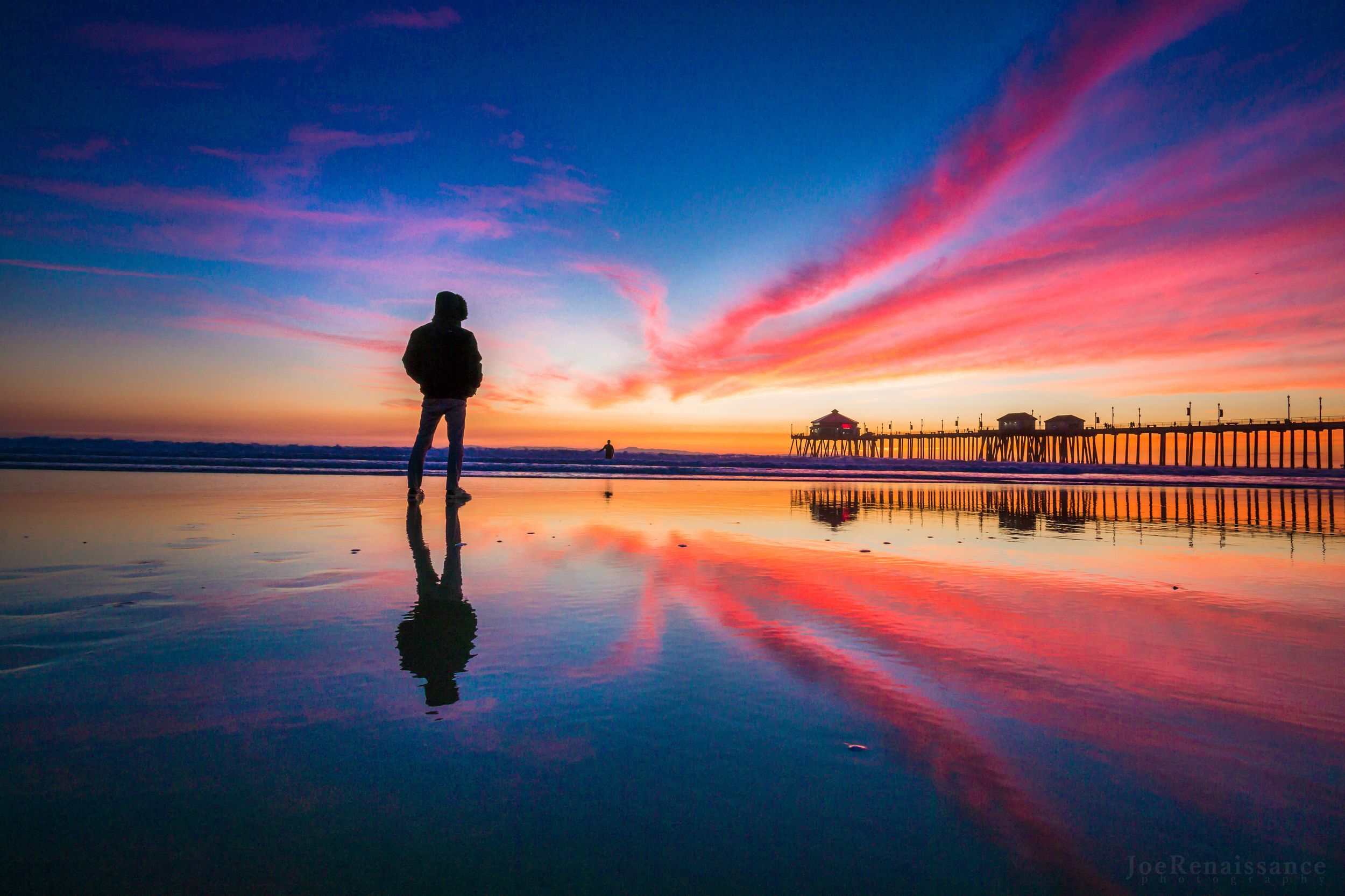 A silhouetted figure stands on wet beach sand at sunset with a pier and vibrant pink clouds reflected in the water.