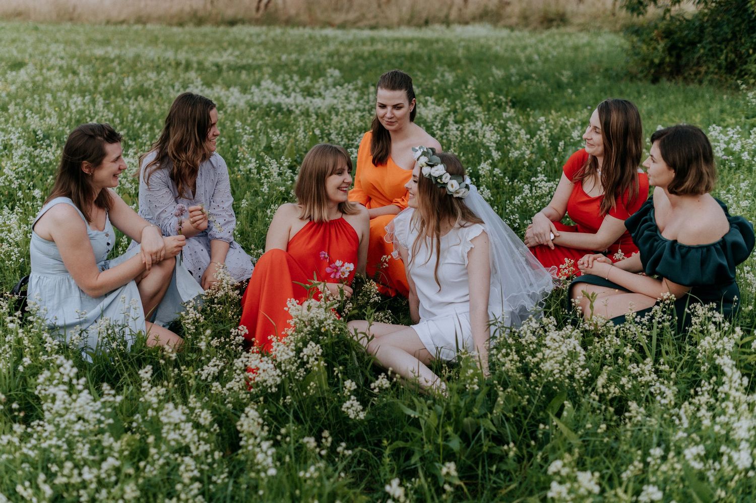 A group sitting together in a blooming wildflower field wearing colorful dresses on a sunny day.