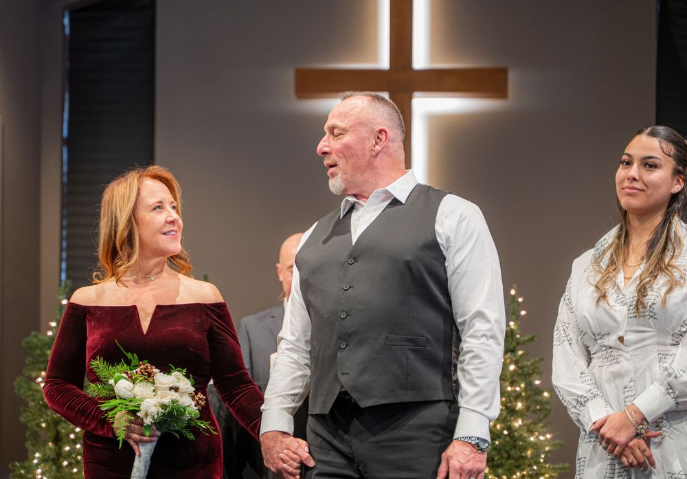 Three people stand before an illuminated cross during a wedding ceremony at a church with twinkle lights in the background.