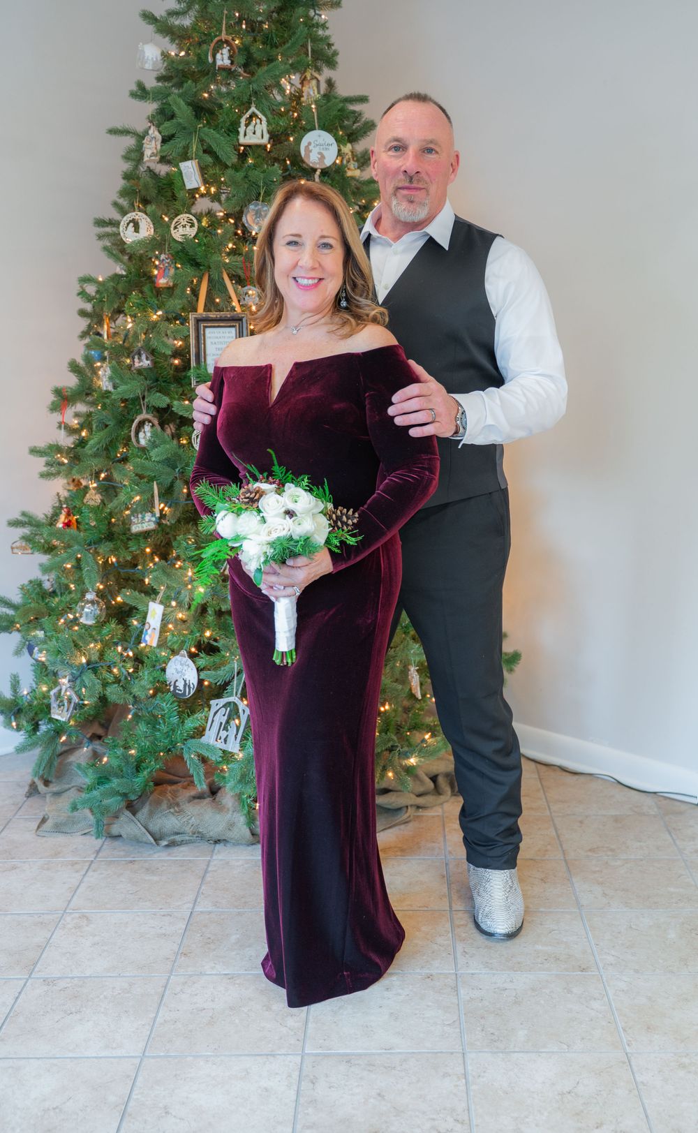 Couple poses by decorated Christmas tree with woman in burgundy velvet gown holding white bouquet.