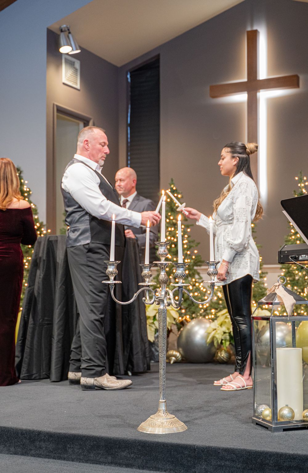 People participate in a worship ceremony on stage with a large illuminated cross and decorative candelabras in a church setting.
