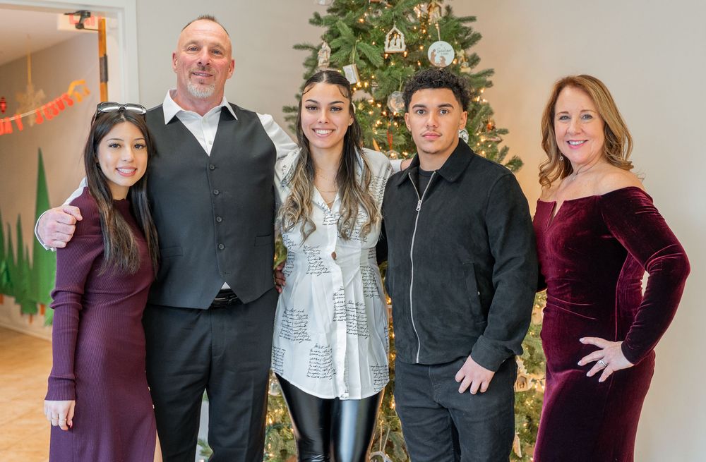 Group of people in casual and formal attire standing together in front of a decorated Christmas tree indoors.