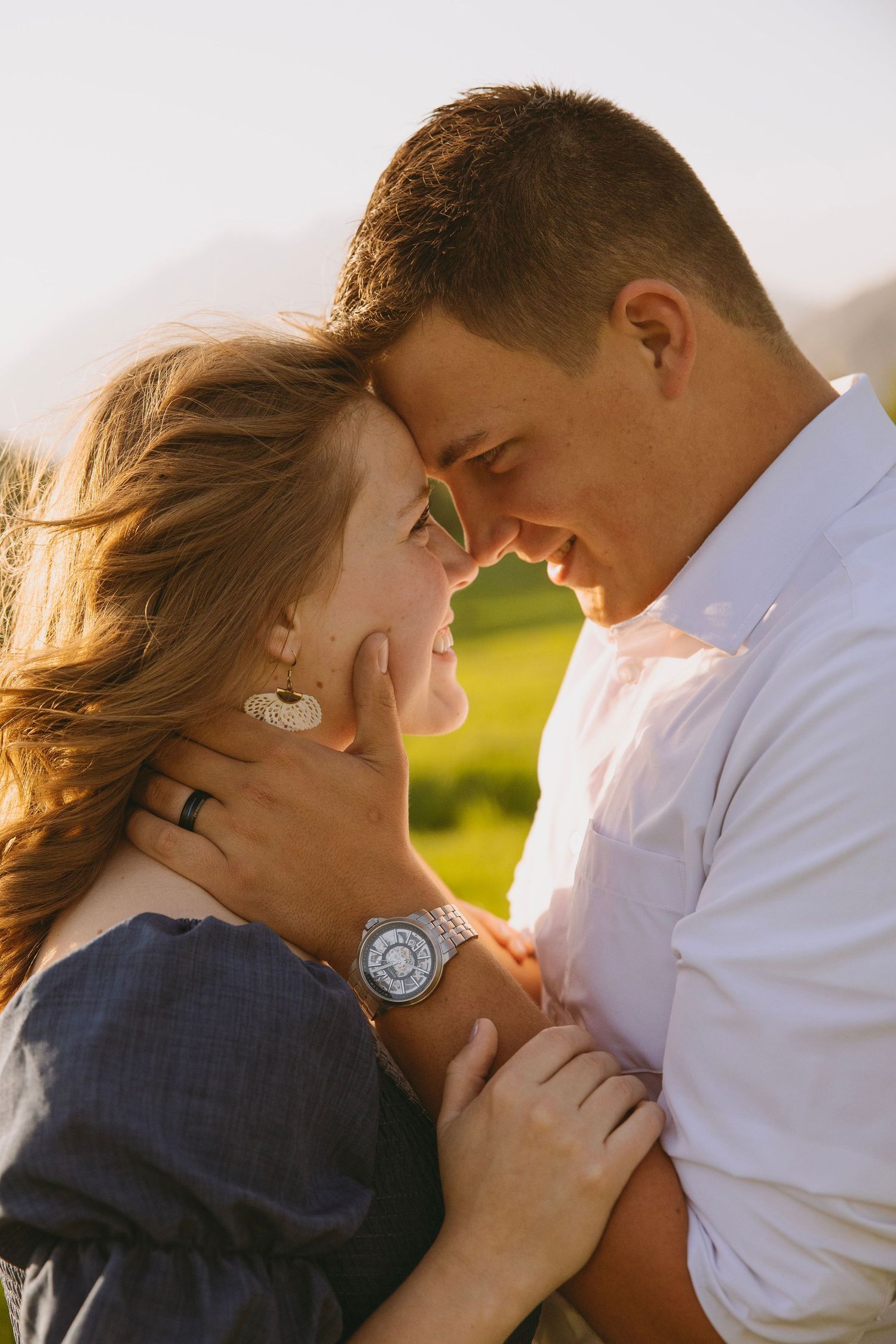Bride in flowing white dress holds bouquet while standing on wooden deck surrounded by evergreen mountains at sunset.