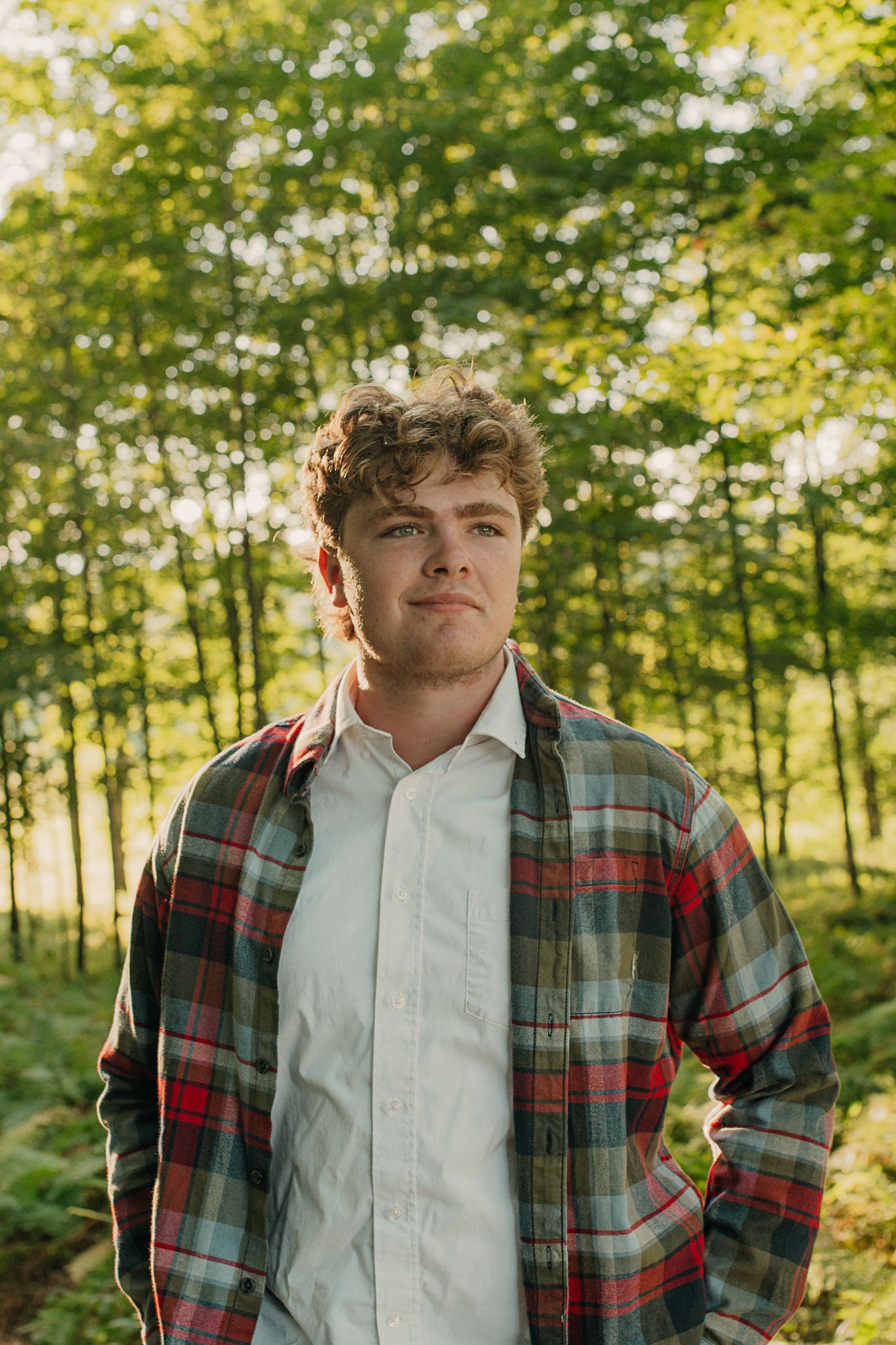 Young person wearing plaid flannel shirt and white button-up in outdoor forest setting with sunlit trees in background.