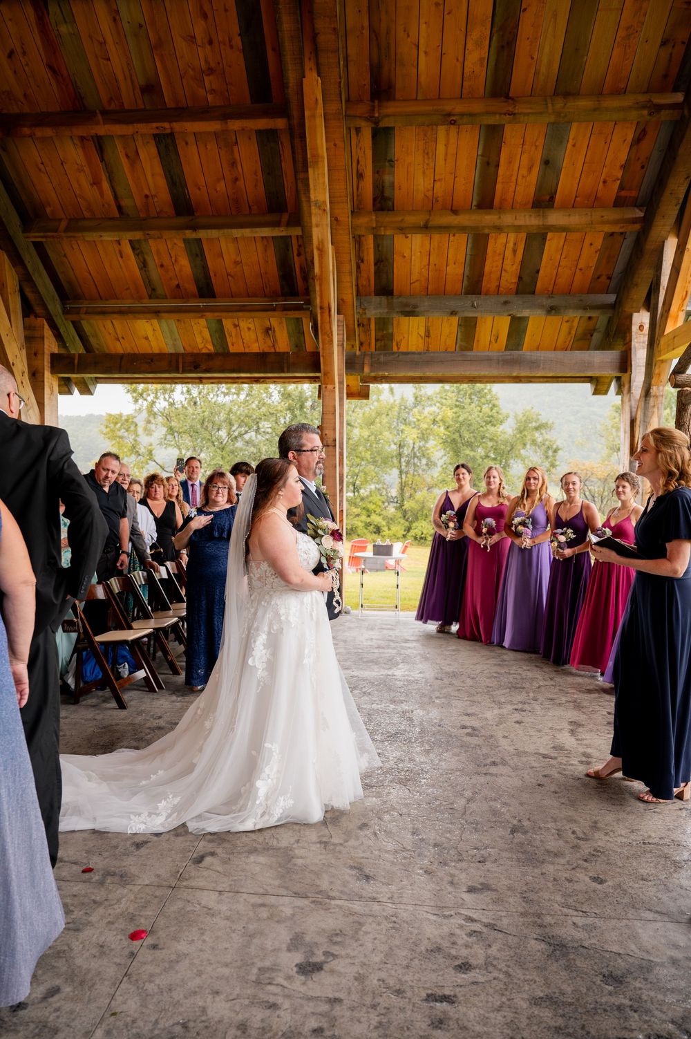 Wedding ceremony guests gather under a rustic wooden pavilion decorated with burgundy and purple accents.