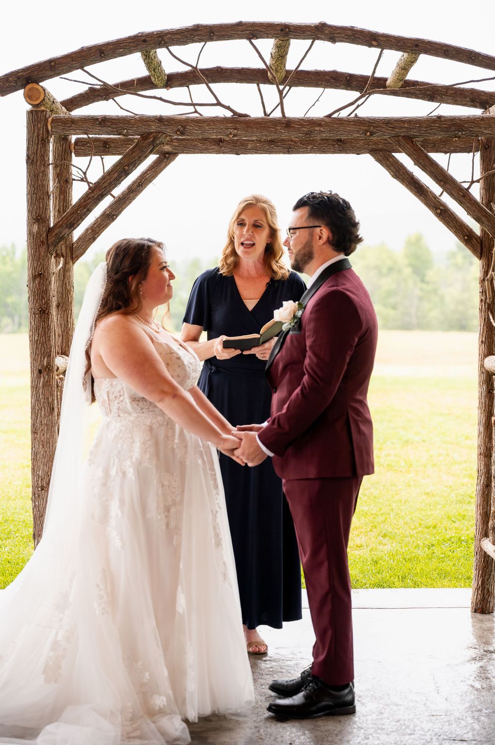 A couple exchanges vows during their wedding ceremony under a rustic wooden pavilion in a countryside setting.