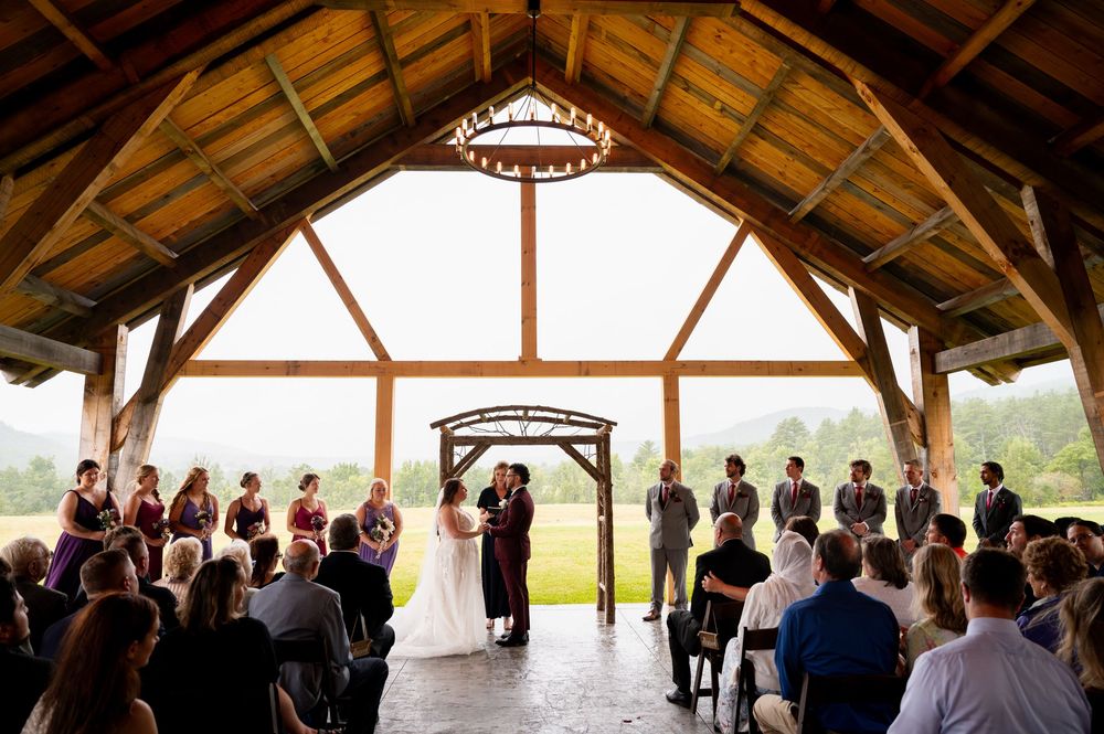 A rustic timber-frame outdoor pavilion hosts an intimate wedding ceremony with guests gathered under wooden beams.