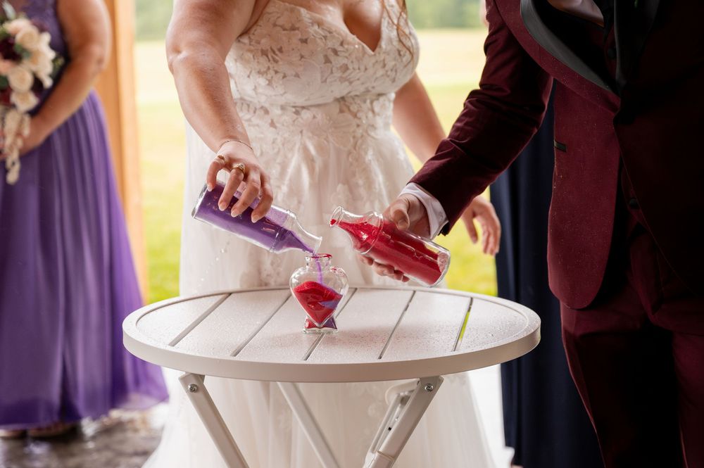 Two people pour red liquids into a glass during a wedding ceremony ritual on a white table outdoors.