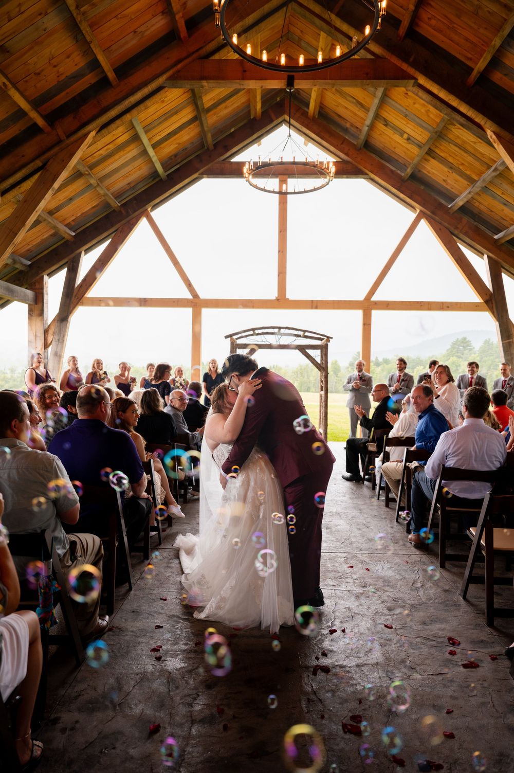 A rustic wooden barn wedding ceremony venue with vaulted ceiling and chandelier while guests look on and bubbles float through air.