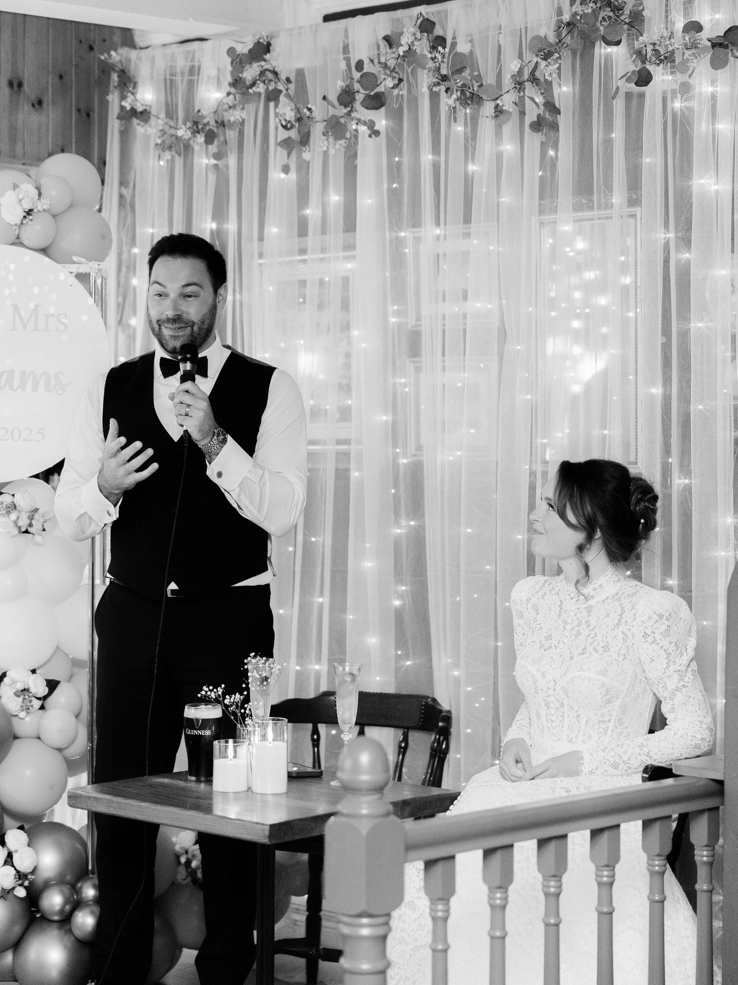 Elegant wedding reception setup with white balloon decor, string lights and floral arrangements against sheer curtains.