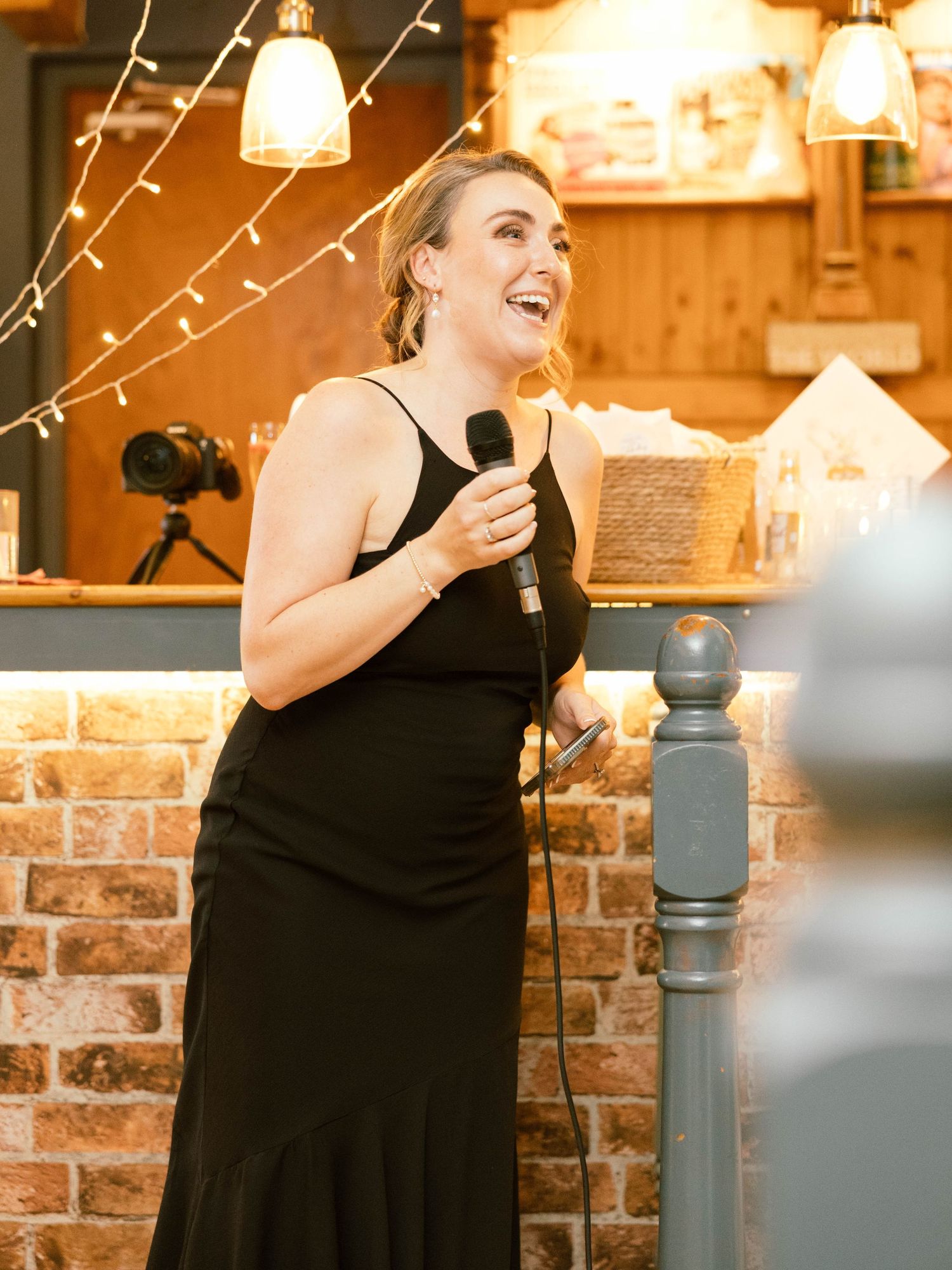 A speaker in a black formal dress gives an animated speech at a rustic venue with exposed brick and string lights.