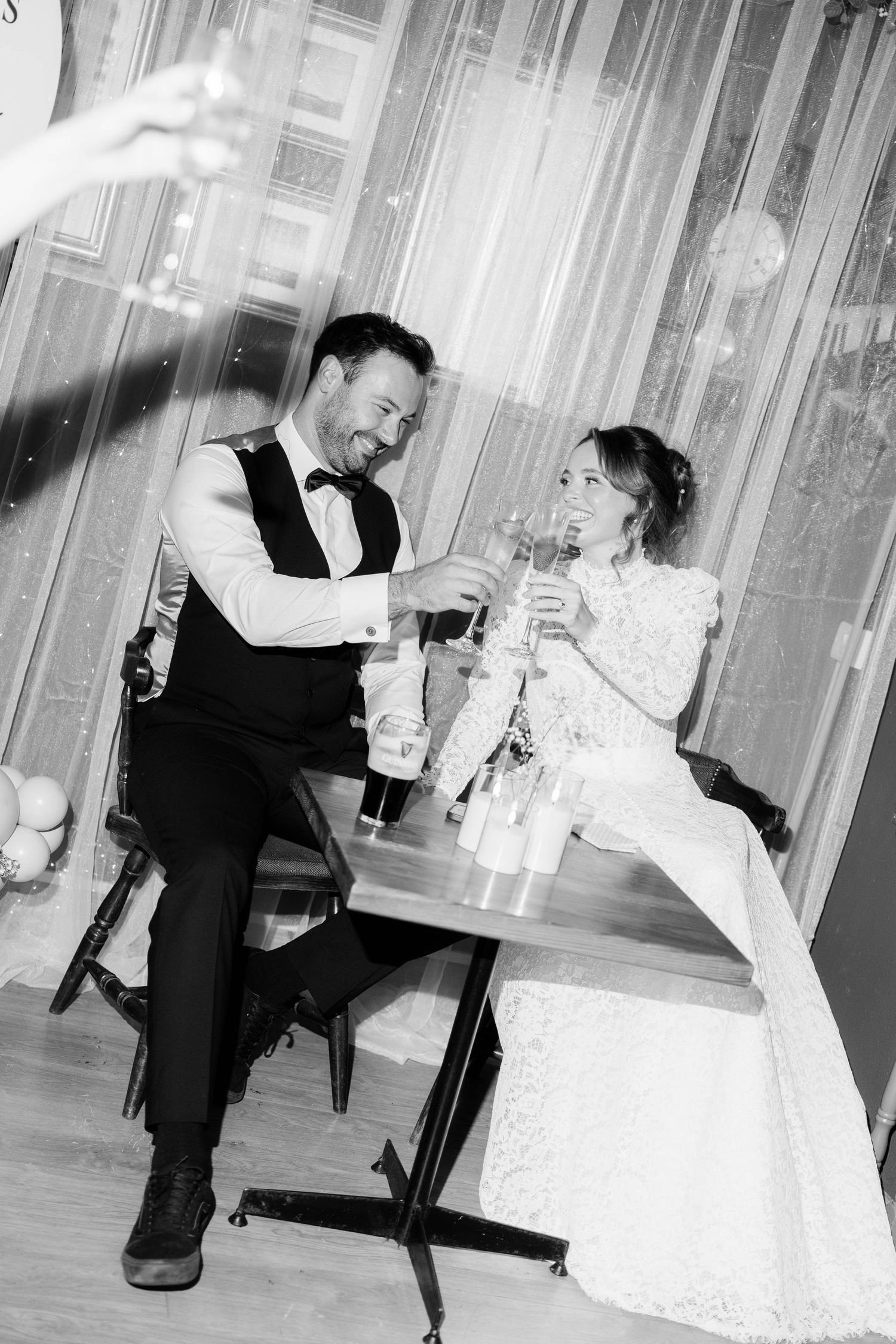 A couple shares a joyful moment with beverages at their wedding reception, captured in black and white photography.
