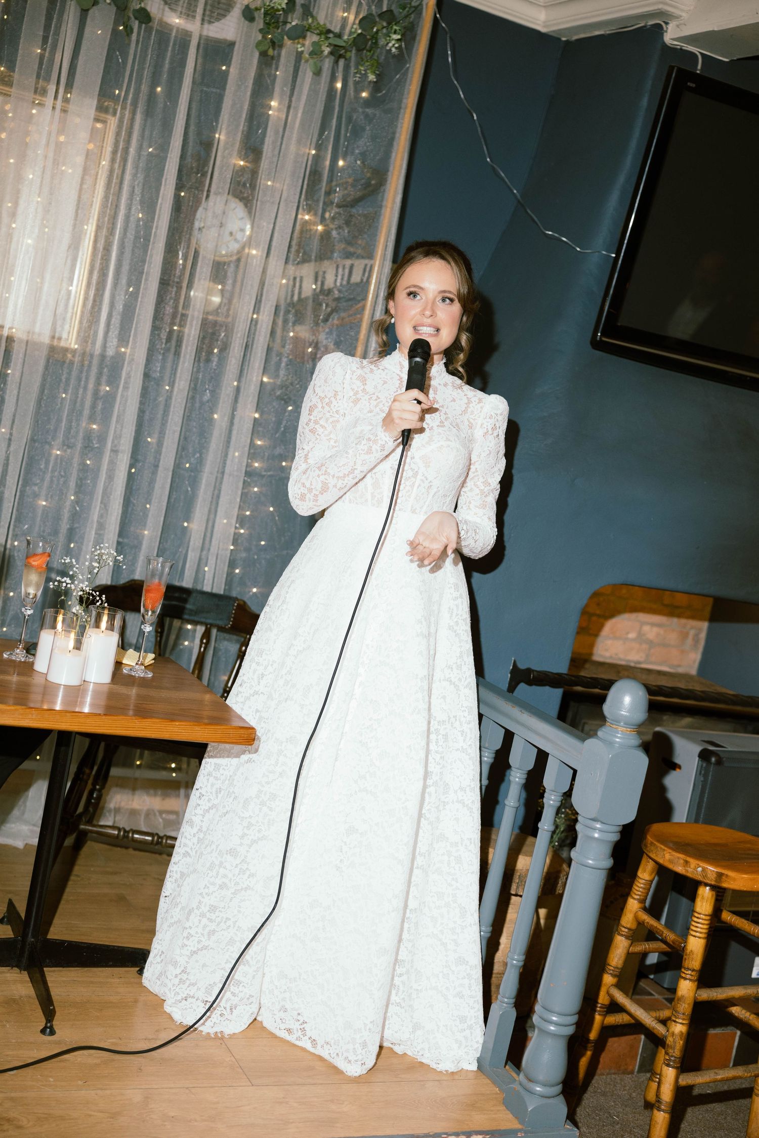 Person in an elegant white lace wedding dress holds a microphone while standing in an indoor venue with sheer curtains.