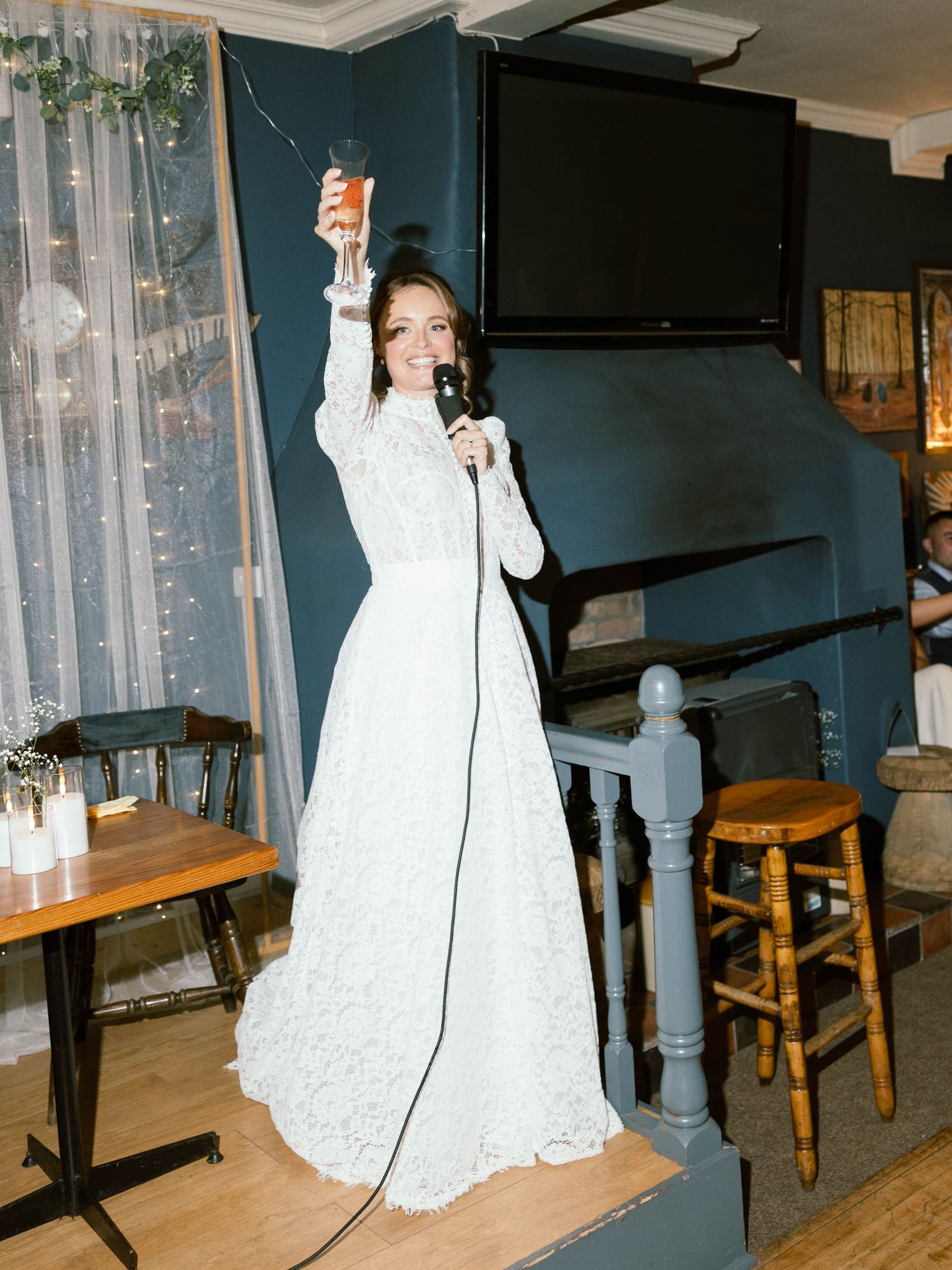 A speaker in a white lace gown raises her hand while speaking into a microphone during an indoor event.