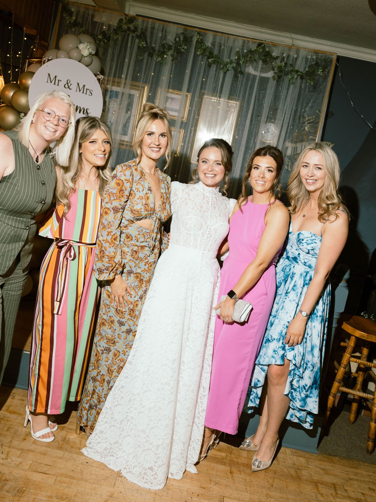 A group of women in colorful dresses posing together at a wedding celebration with decorative lighting and Mr & Mrs sign.